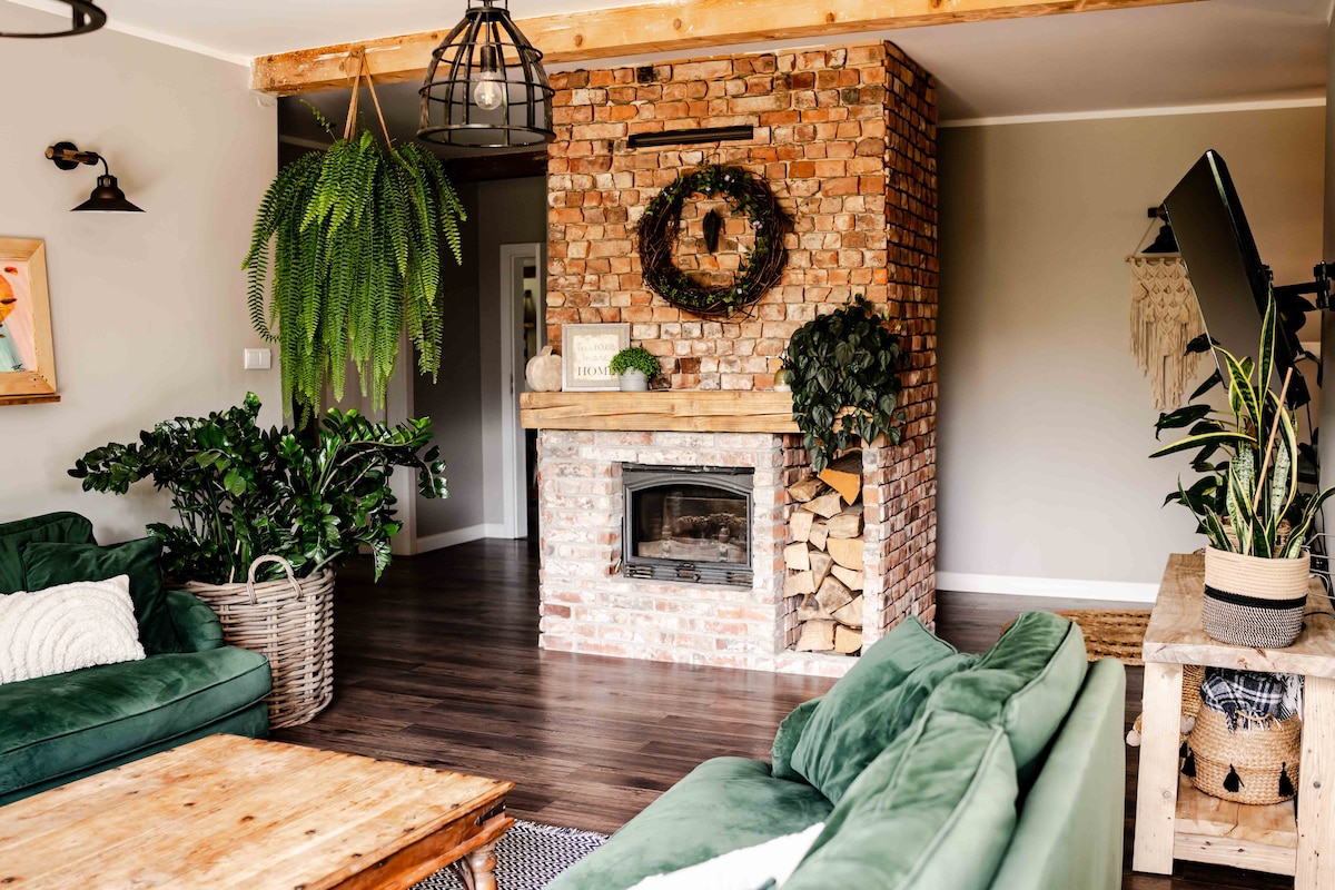 The living area features a sturdy brick fireplace surrounded by wooden accents. Cozy green sofas are arranged around a wooden coffee table. Abundant greenery from potted plants enhances the space, while natural light fills the room through nearby windows.