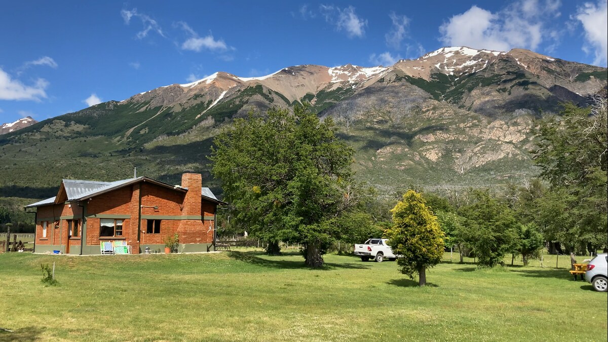A brick cabin is set against a backdrop of majestic snow-capped mountains. Lush green grass surrounds the property, along with scattered trees. A single car is parked nearby, emphasizing the peaceful, rural environment.