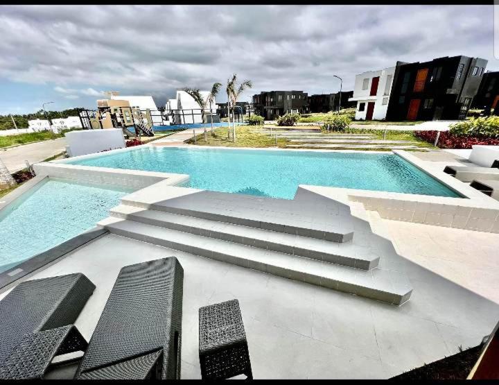 A spacious outdoor area features a glimmering swimming pool surrounded by modern villas. Steps lead into the pool, while a shallow area is visible for relaxing. Comfortable outdoor seating is arranged nearby, providing a serene space to enjoy the scenery.