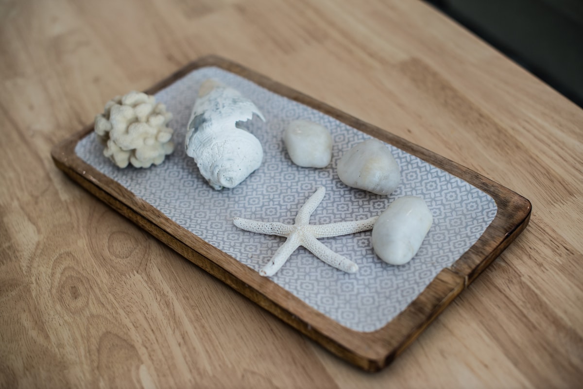 A wooden tray is displayed on a light wooden surface, containing several seaside-themed decorative items. Featured are a starfish, smooth white stones, and assorted seashells, all arranged on a textured linen cloth.