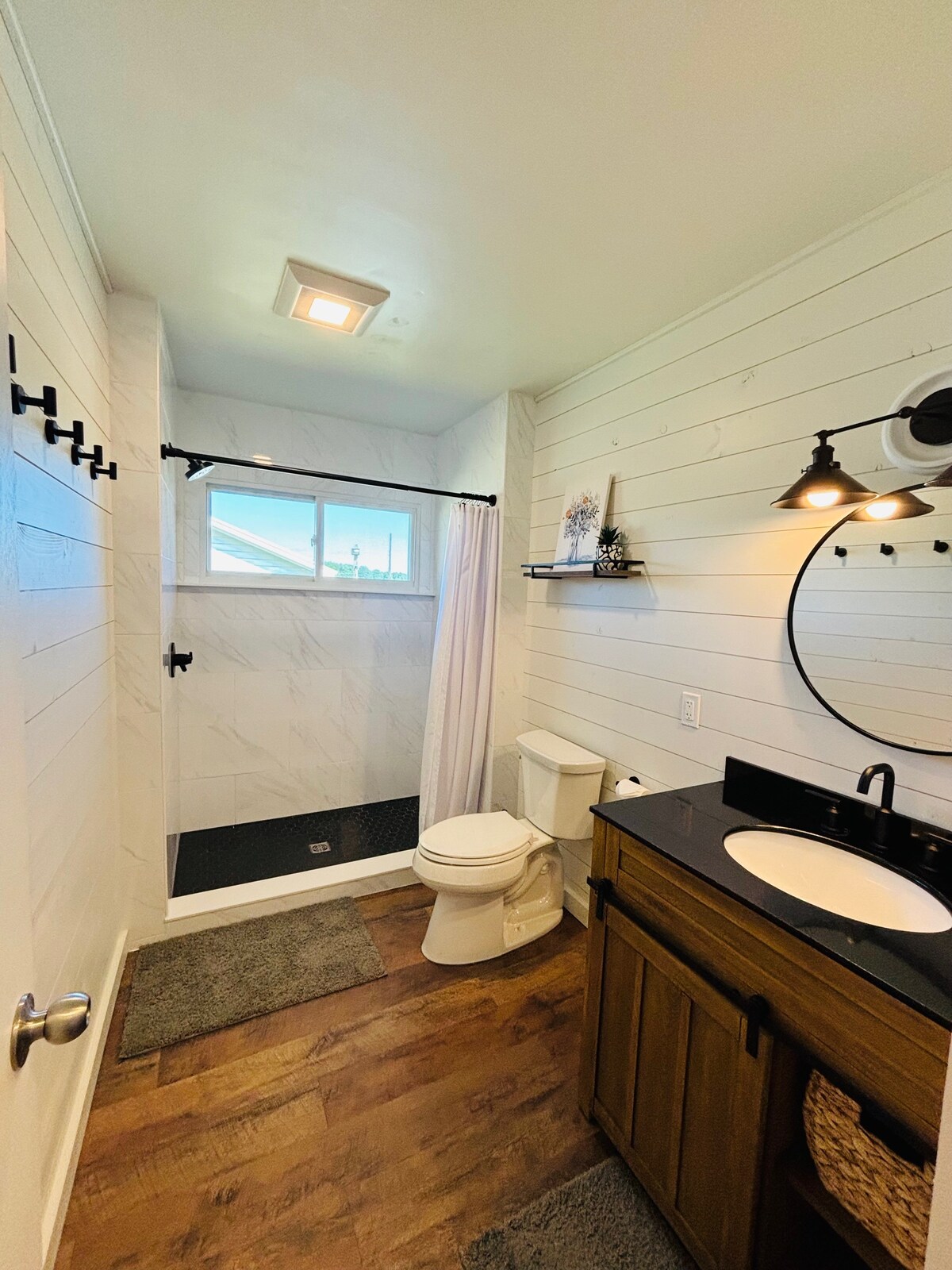 A bright bathroom features a walk-in shower with a clear glass door, white shiplap walls, and a large mirror above a dark countertop sink. A toilet is positioned adjacent to the shower, while two bath mats provide comfort underfoot.