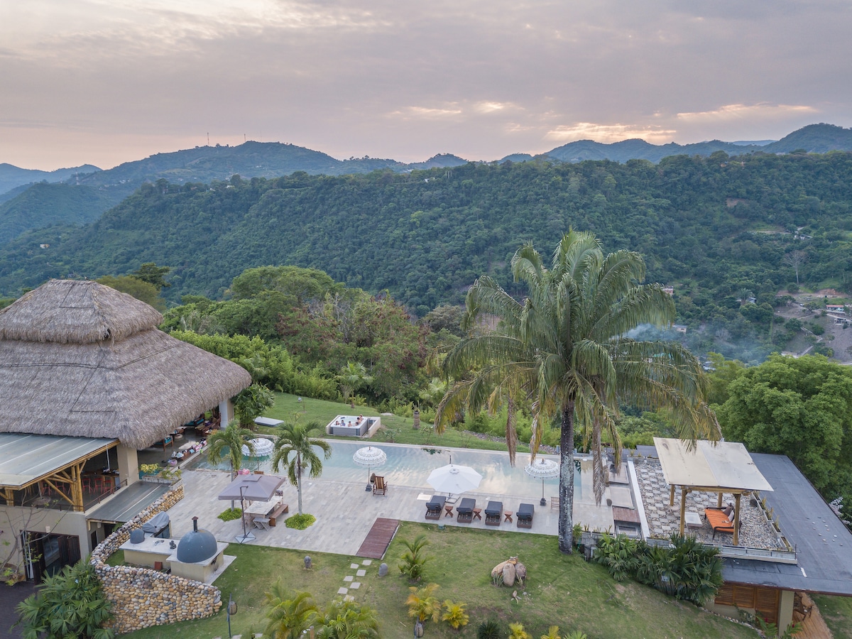 An aerial view showcases a spacious outdoor area featuring a swimming pool with sun loungers. Lush greenery surrounds the landscape, complemented by a thatched-roof structure. Mountains are visible in the background, with a soft sky tinted by sunset hues.