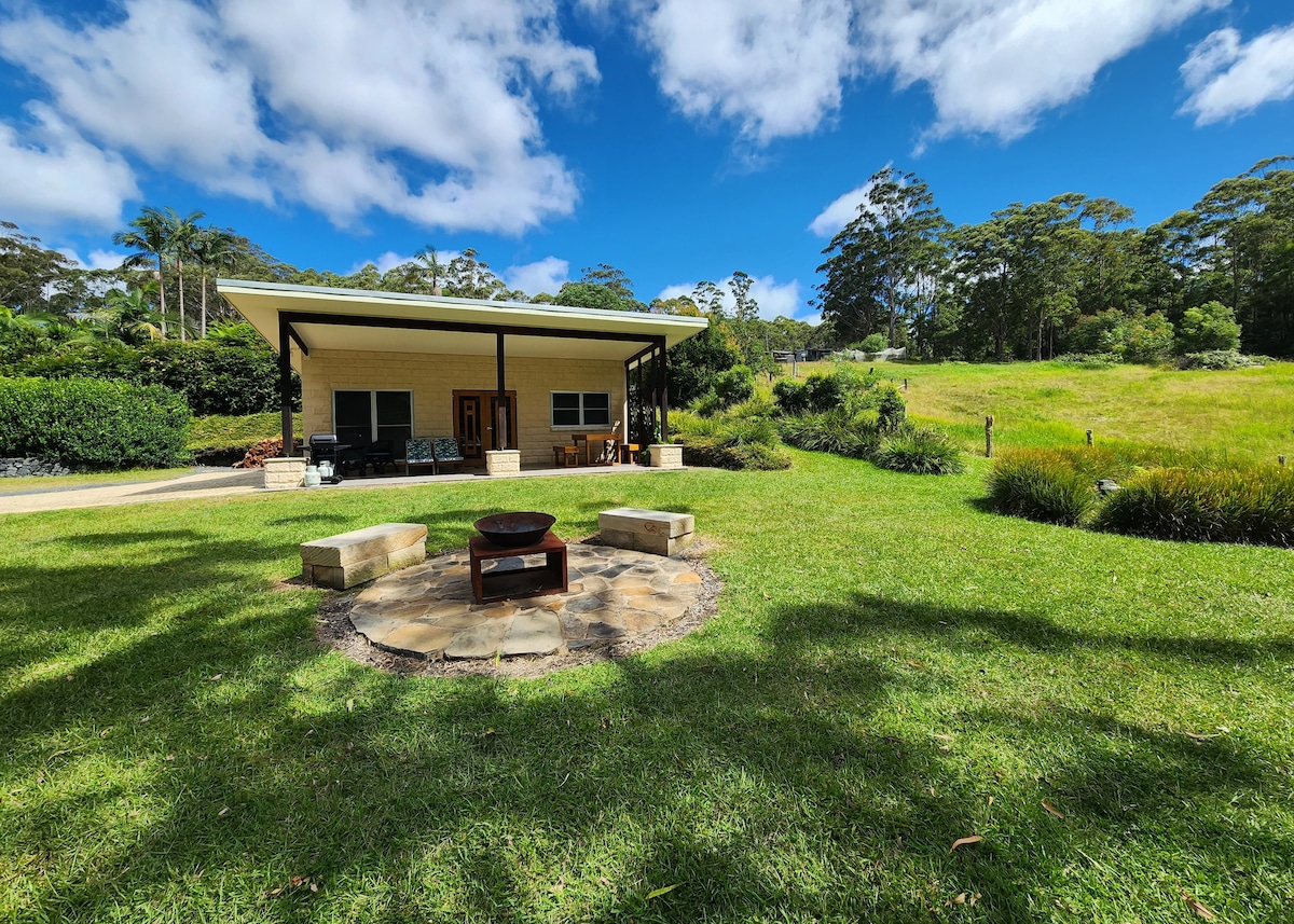A spacious cabin is set against a lush landscape, featuring a well-maintained lawn and trees in the background. A circular stone fire pit is arranged in front of the cabin, providing a natural gathering space. Soft clouds drift in the bright blue sky above.