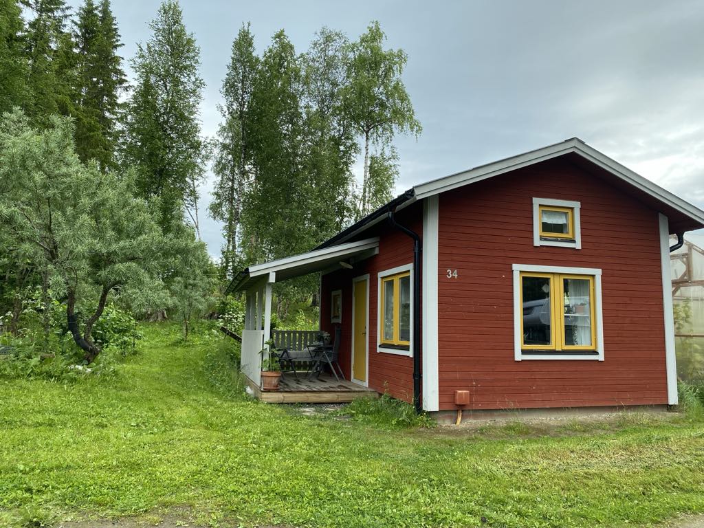 A charming red cottage is surrounded by lush greenery, featuring a covered porch with a seating area. The exterior showcases large windows with yellow frames, inviting natural light into the home. A tranquil atmosphere is present with trees and open grass nearby.