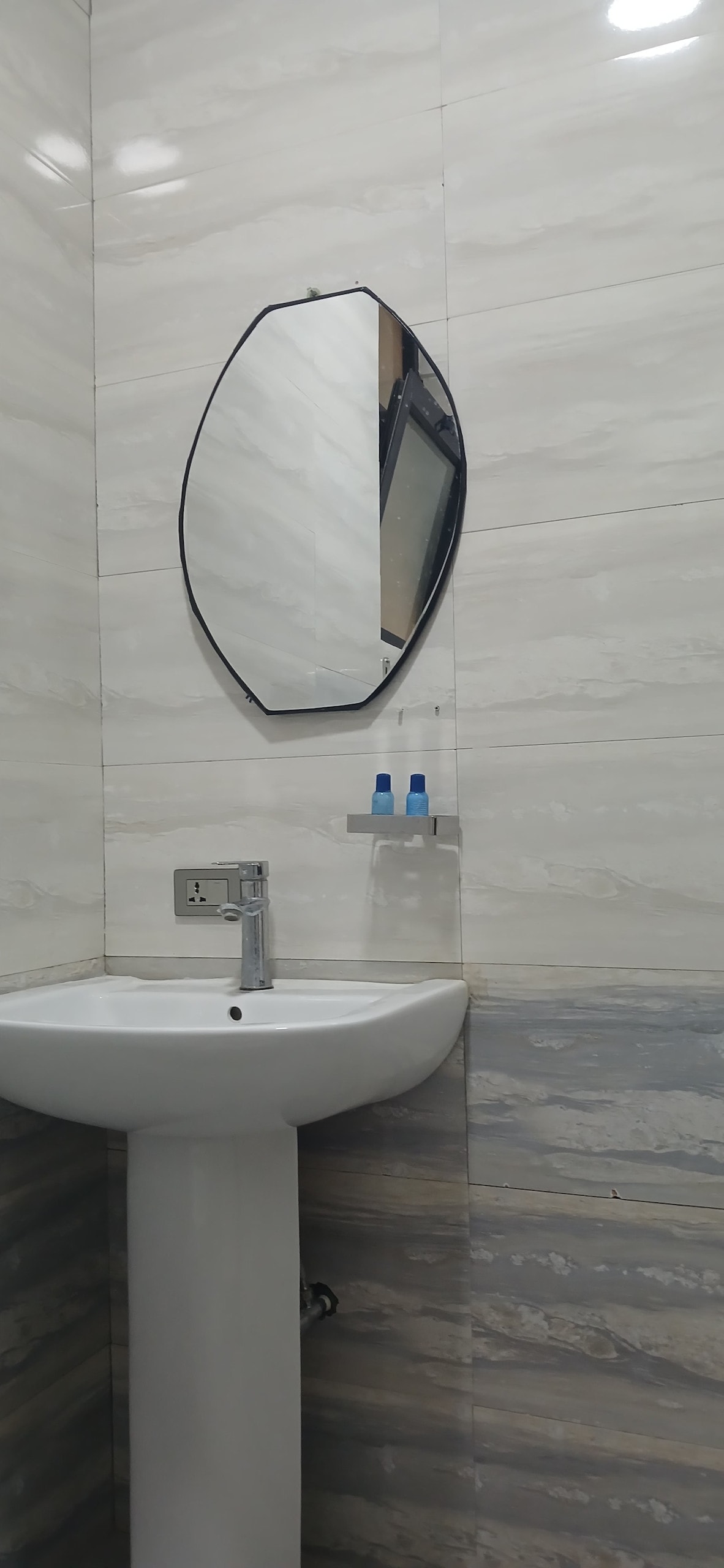 A modern bathroom features a sleek oval mirror above a compact sink. The walls are adorned with elegant light grey tiles, while a contrasting darker marble pattern is present at the base. A small shelf holds two blue bottles, adding a subtle touch of color.
