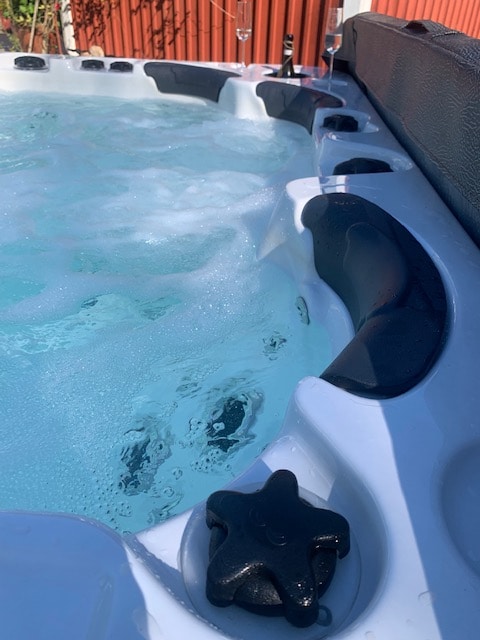 A close-up view of a hot tub featuring bubbling water, surrounded by ergonomic seating. The structure showcases a textured rim and control knobs. Soft sunlight highlights the clear water while the background includes a hint of a red wooden wall.