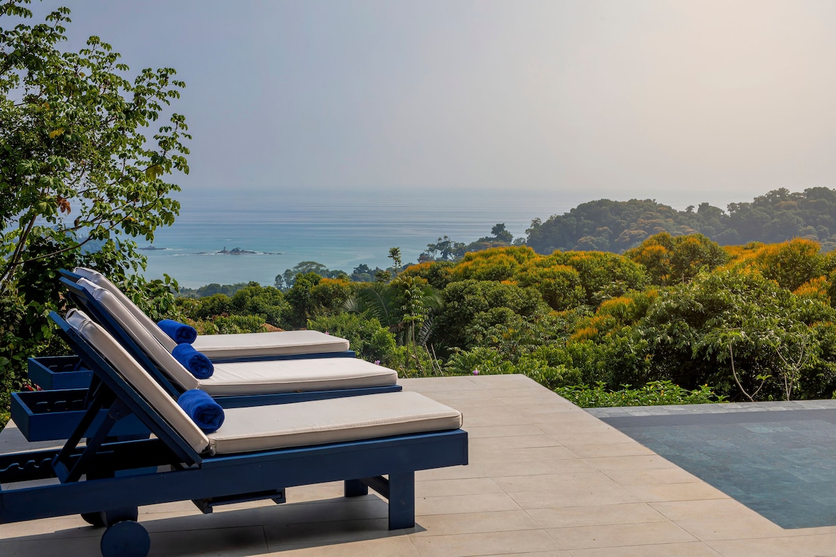 Sun loungers with rolled towels are positioned near the infinity pool, overlooking a panoramic view of the ocean. Lush greenery and vibrant trees create a natural backdrop, while a hazy horizon blends sky and sea in soft tones.
