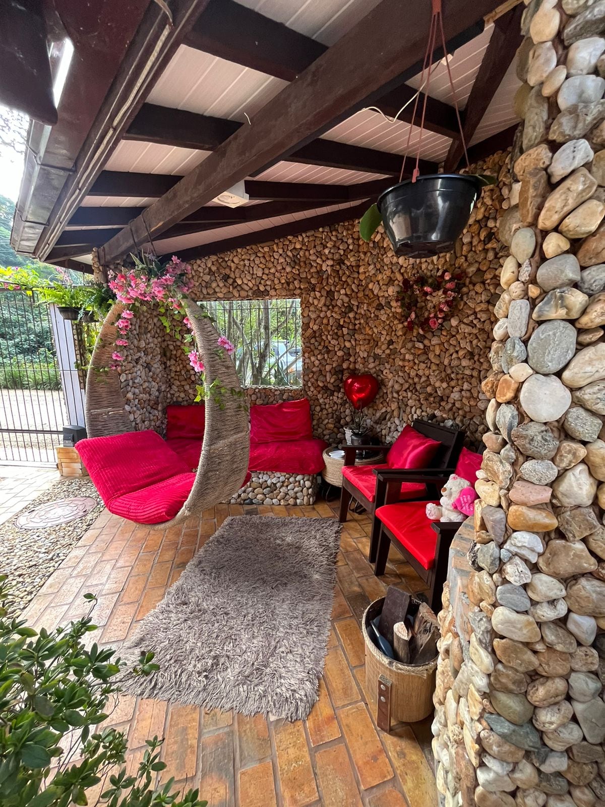 A cozy outdoor seating area is surrounded by a stone wall, featuring a hanging swing adorned with pink cushions. Two red chairs complement the space, which is accented by vibrant floral decorations. A textured rug lies on the brick floor, adding warmth to the environment.