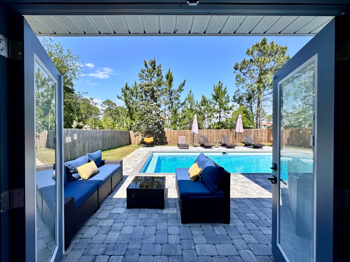 Double French doors open to a serene outdoor space featuring a private saltwater pool. Lounge chairs with plush cushions surround the pool, while sun umbrellas provide shade. The well-maintained backyard includes a wooden fence and lush greenery, creating a tranquil environment.