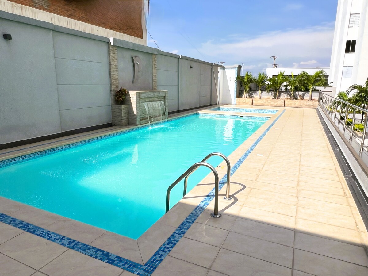 The swimming pool is surrounded by a tiled terrace, featuring a gentle waterfall feature. Sunlight reflects off the clear water, and greenery is visible in the background. The area provides a spacious setting for relaxation and leisure.
