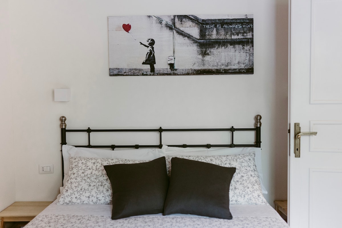 A cozy bedroom is highlighted by a metal bed frame adorned with white linens and two dark decorative pillows. Above the bed, a wall art piece depicts a child releasing a heart-shaped balloon against a textured background, adding a touch of charm to the space.