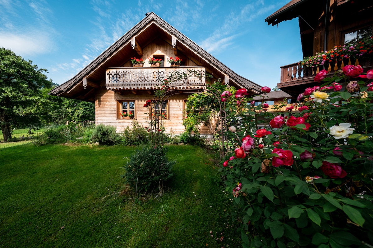 A charming wooden house is set against a blue sky, featuring a balcony adorned with colorful flowers. Lush greenery surrounds the property, and vibrant rose bushes bloom in the foreground, creating a serene outdoor setting.