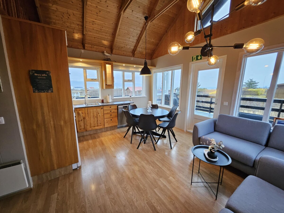 An open living space showcases a modern kitchen and a dining area featuring a round table surrounded by black chairs. Large windows allow natural light to fill the room, highlighting wooden accents and a cozy seating area with a grey sofa.