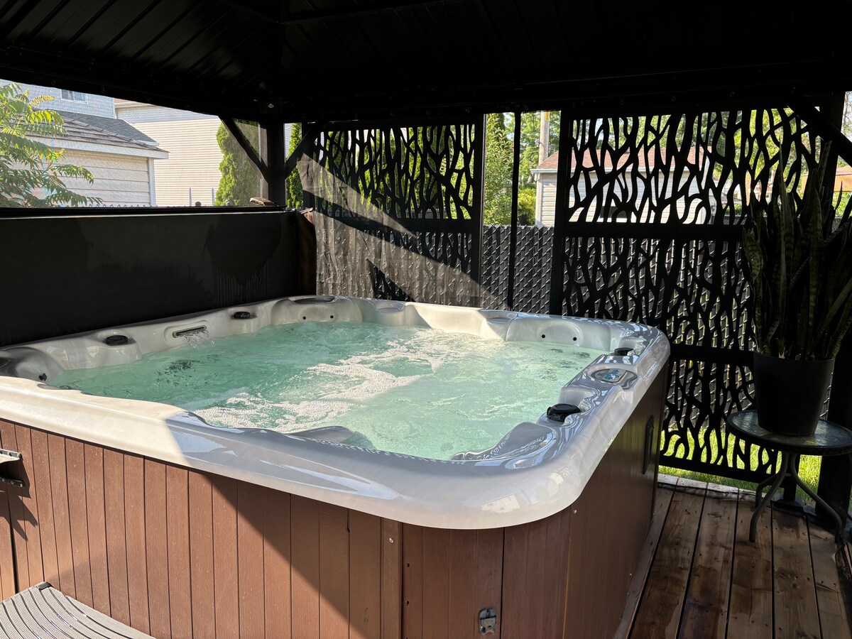 A hot tub is positioned under a shaded structure, surrounded by a wooden deck. Intricate privacy screens provide seclusion, while natural light filters through the openings. Green water bubbles gently, inviting relaxation in a peaceful outdoor setting.