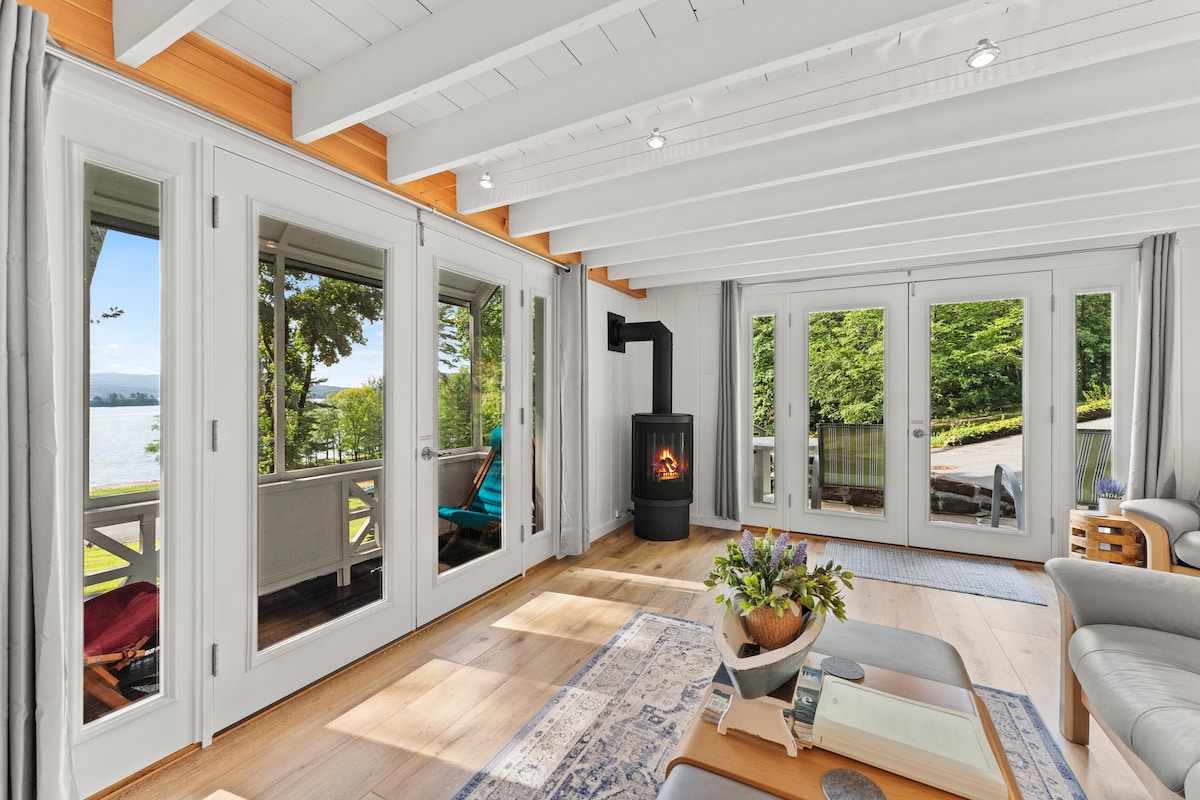 A bright living area is shown with a gas stove positioned near large glass doors that offer lake views. Natural wood beams enhance the ceiling. A light-colored couch and a decorative plant center the space, complemented by a soft area rug.