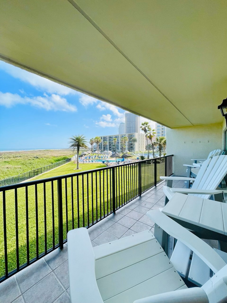 A balcony view showcases white adirondack chairs overlooking a grassy area and swimming pool. Palm trees are seen swaying in the gentle breeze, framed by a clear blue sky and ocean in the distance. The condo's peaceful surroundings are accentuated by a sense of tranquility.