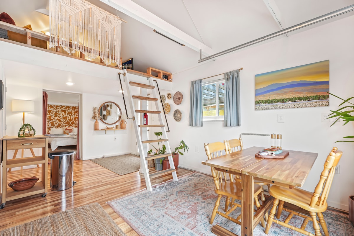 An open living area features warm wooden floors and rustic furniture. A dining table with four chairs is positioned beside a large window. A ladder leads to a lofted sleeping area, while an art piece adorns the wall with calming landscape tones.