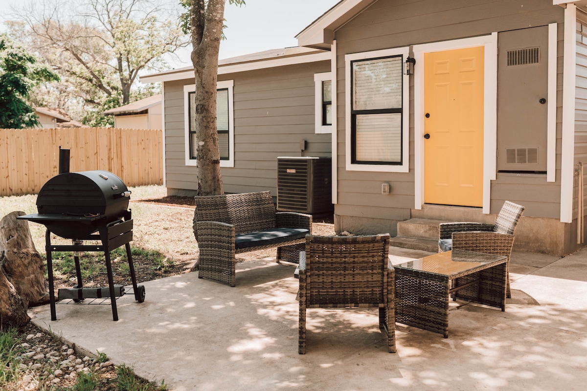 An outdoor patio features a BBQ smoker alongside four wicker chairs and a glass-topped table. A bright yellow door adds a welcoming touch to the home's exterior, which is framed by trees and a wooden fence, creating a comfortable and private atmosphere.