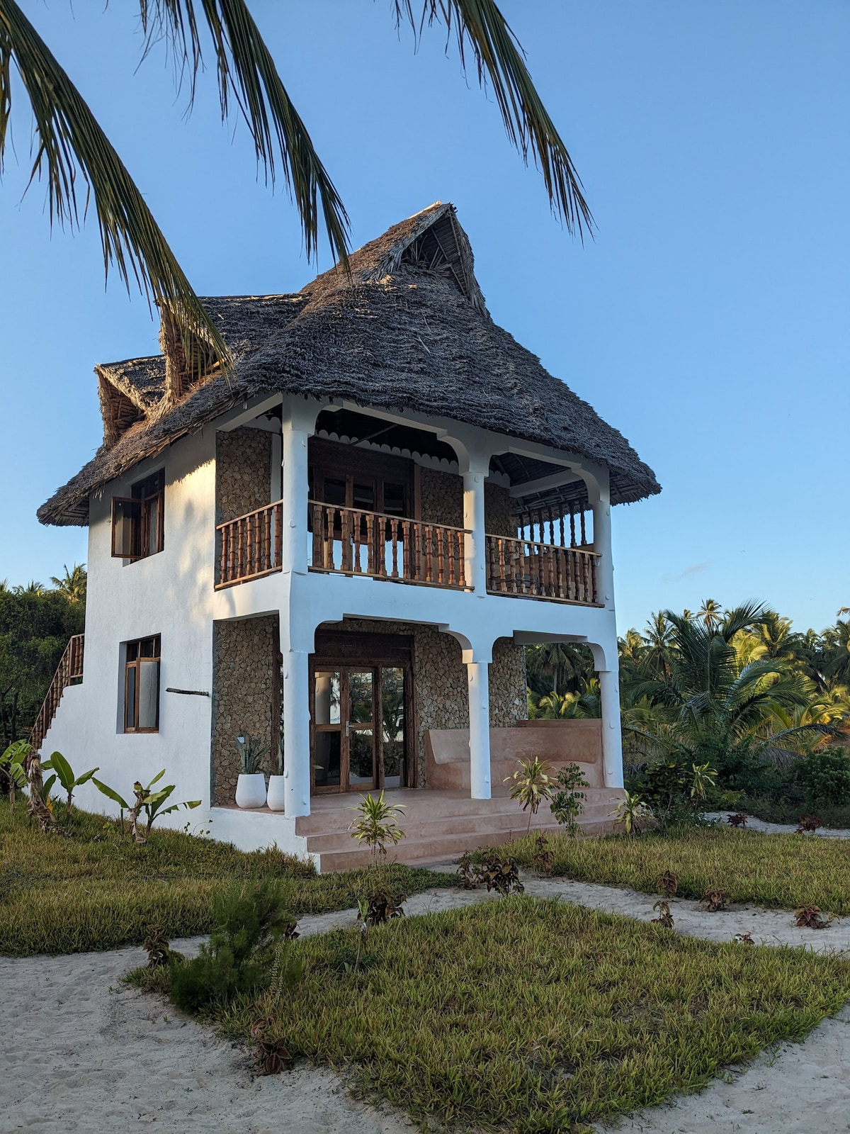 A two-story beach house features a thatched roof and wooden balconies, surrounded by lush greenery and palm trees. The entrance is marked by large glass doors, inviting natural light. The sandy landscape leads to the beautiful beachfront, creating a serene coastal environment.