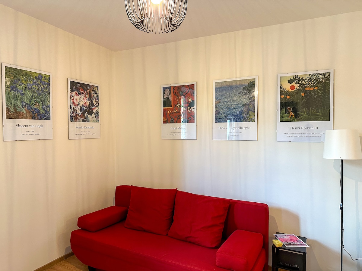 A red sofa is positioned against a light-colored wall adorned with five framed art prints. The prints feature various styles, including works inspired by Van Gogh and other artists. A floor lamp stands nearby, providing additional lighting.