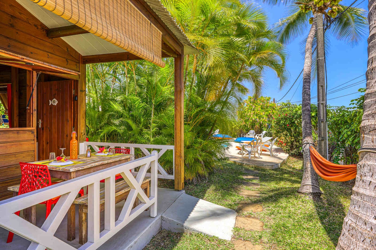 A wooden bungalow is situated amidst lush tropical greenery, featuring a dining table with bright red chairs on the porch. Nearby, a sunny pool area is visible, with lounge chairs arranged for relaxation. A hammock can be seen suspended between two palm trees.