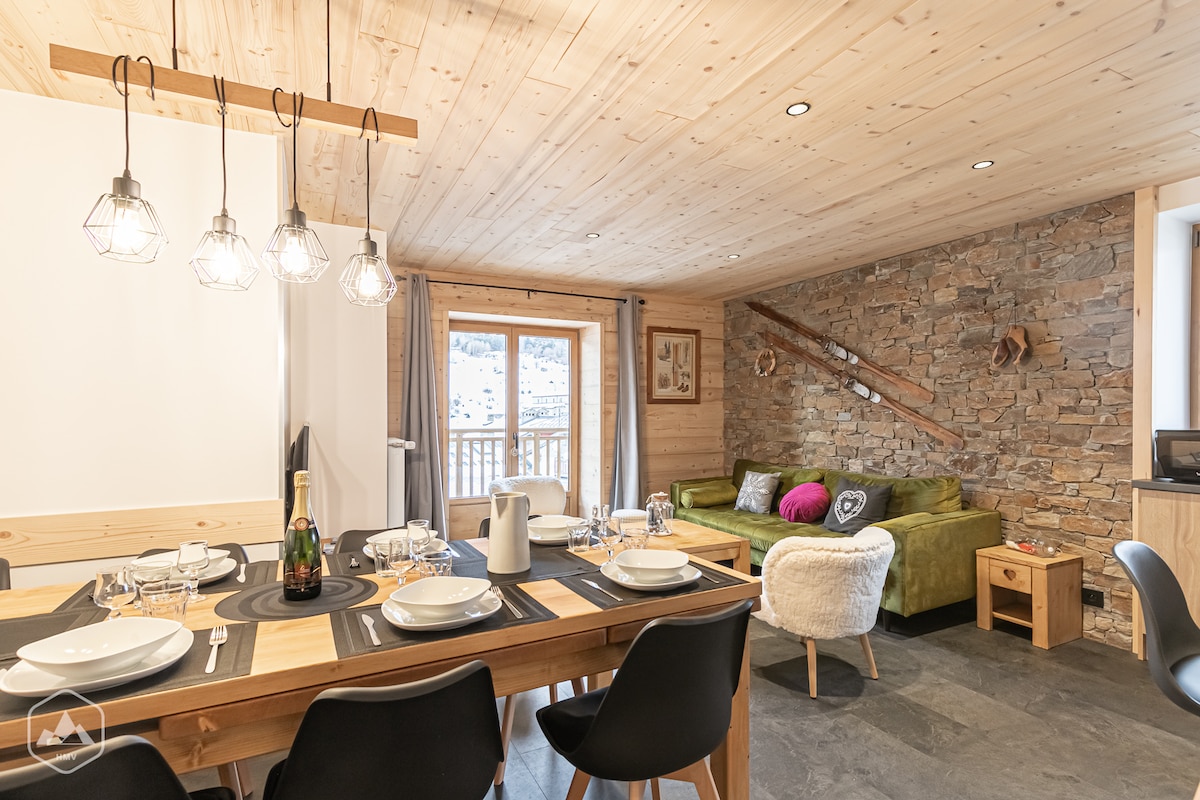 A spacious dining area features a wooden table set for guests, surrounded by modern black chairs. A green sofa and side table are positioned against a stone wall. Warm light is provided by hanging fixtures, and a large window allows natural light, showcasing the outdoor view.