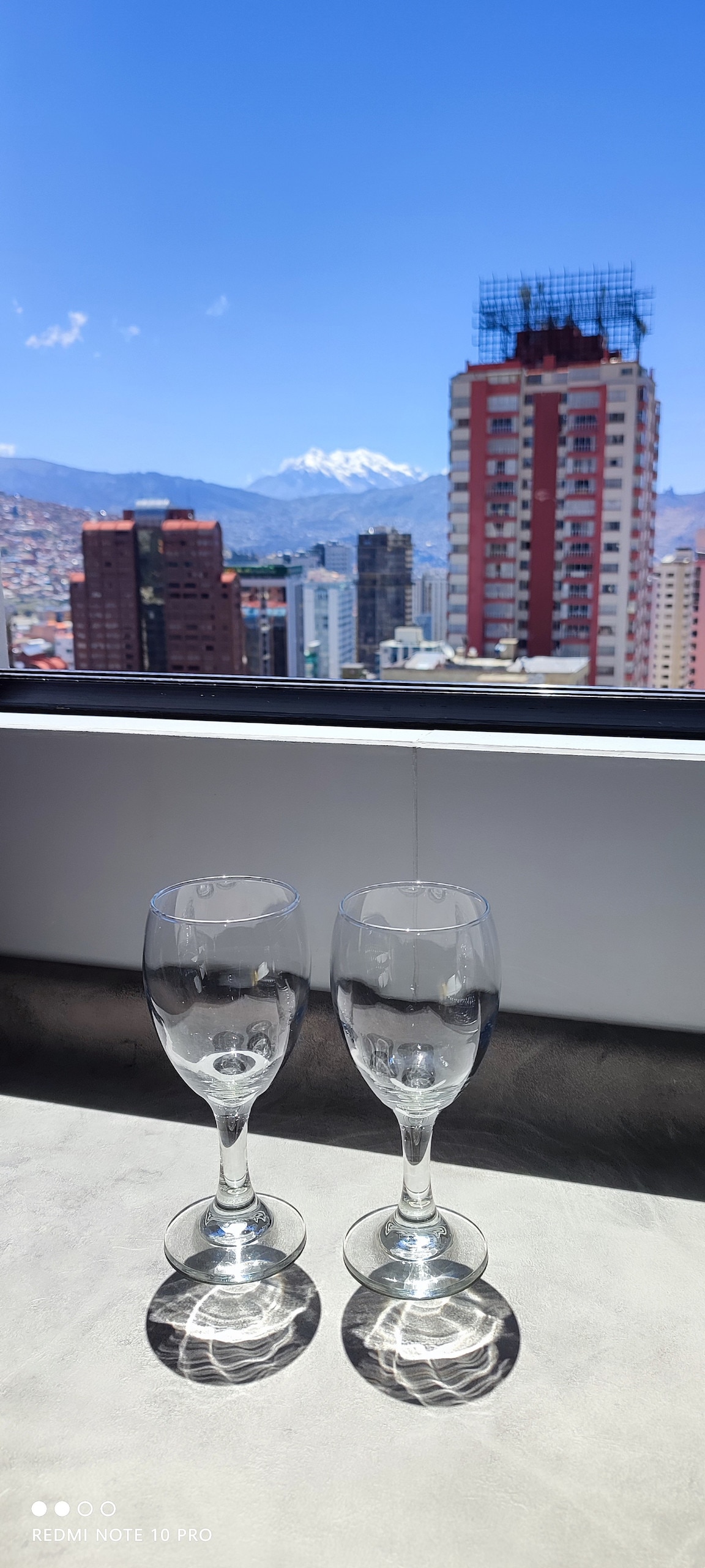 Two elegant wine glasses are placed on a surface, positioned in front of a large window. The view captures a city landscape with buildings and mountains visible in the background, including a peak that may represent Illimani under a clear blue sky.