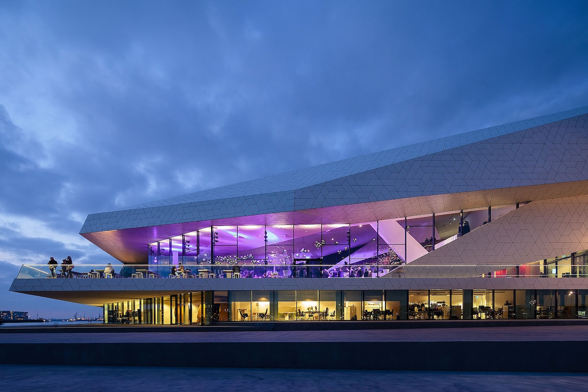 A modern building, characterized by angular architecture and large glass facades, is illuminated in shades of pink and purple at dusk. Outdoor seating is visible on a terrace, suggesting a lively atmosphere. The surrounding area is dark, enhancing the building's striking design.