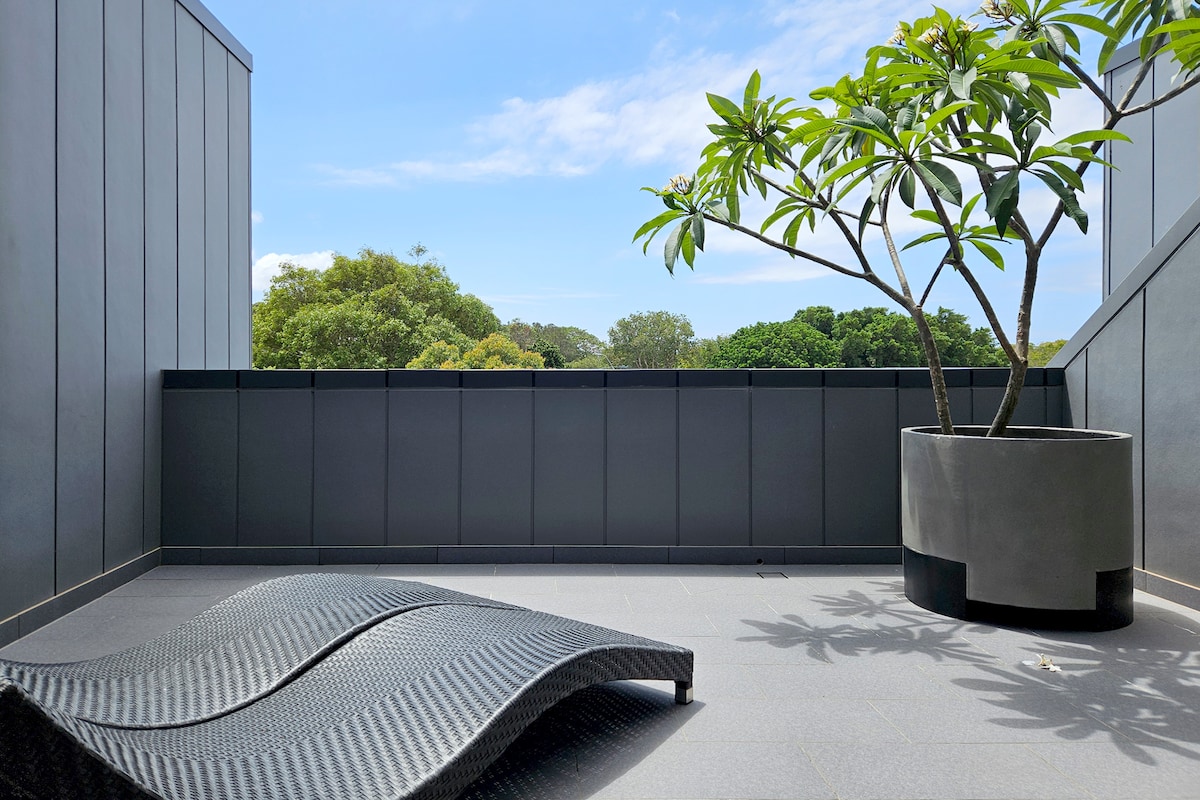A private outdoor space is highlighted by a black lounge chair positioned next to a large potted plant. The area offers a clear view of greenery in the distance, complemented by a bright blue sky.