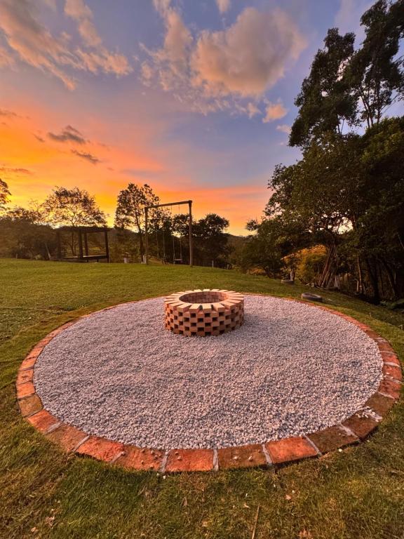 A circular firepit area is surrounded by compacted gravel and bordered by red bricks, situated on a grassy landscape. The sunset creates a warm glow in the sky, while trees are visible in the background, enhancing the outdoor setting.