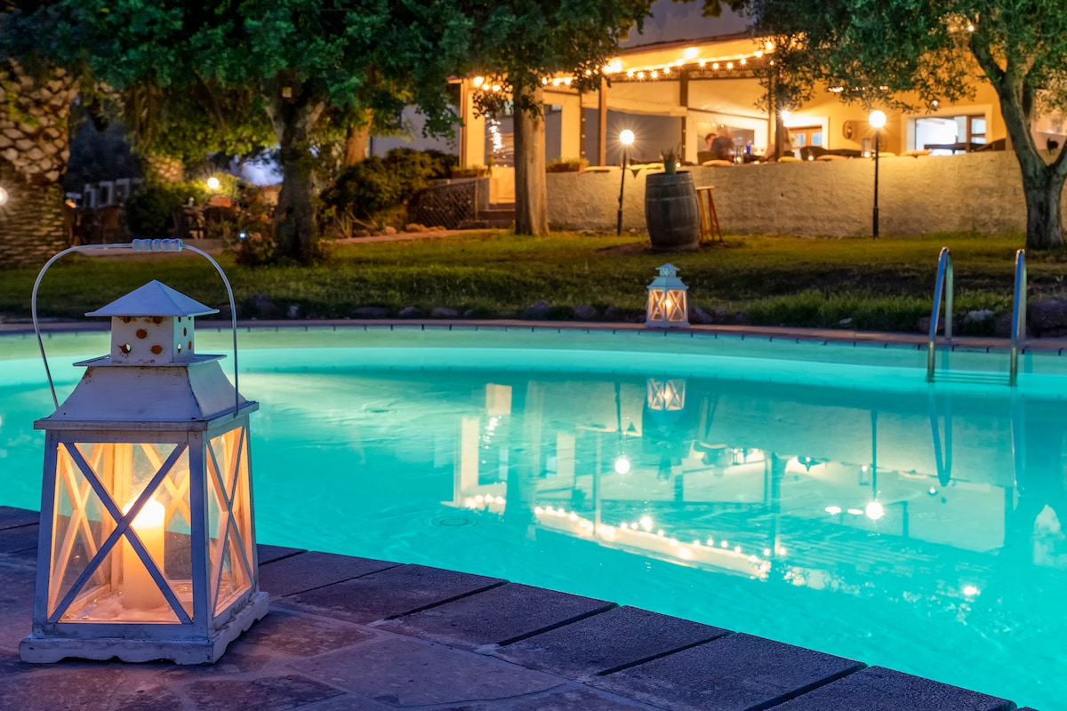 A serene pool area is softly illuminated by lanterns, casting warm reflections on the tranquil water. In the background, the welcoming restaurant area is visible, enhancing the peaceful atmosphere of the outdoor space.
