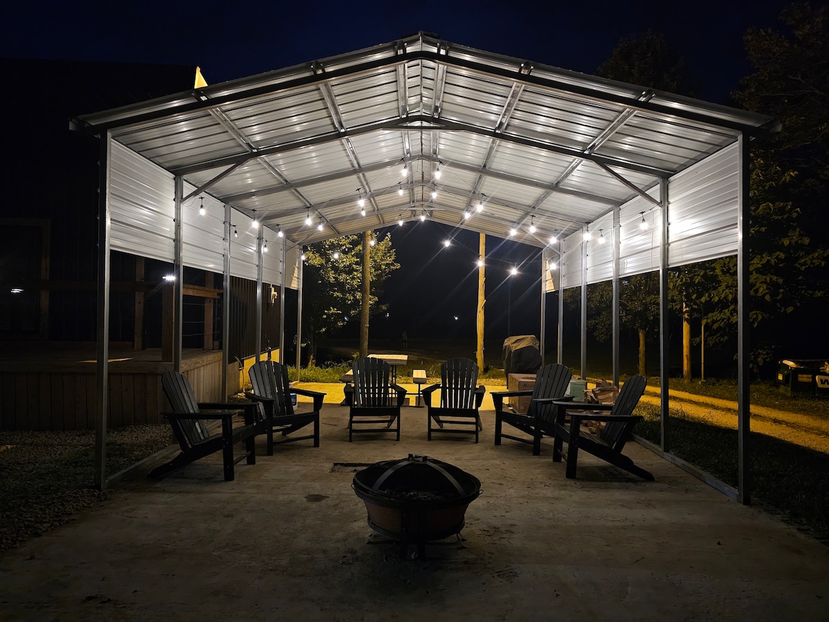 An open-air pavilion is illuminated by string lights, creating a welcoming gathering space. Several Adirondack chairs are arranged around a central fire pit, inviting relaxation. The surrounding area features trees and pathways, enhancing the connection to nature.