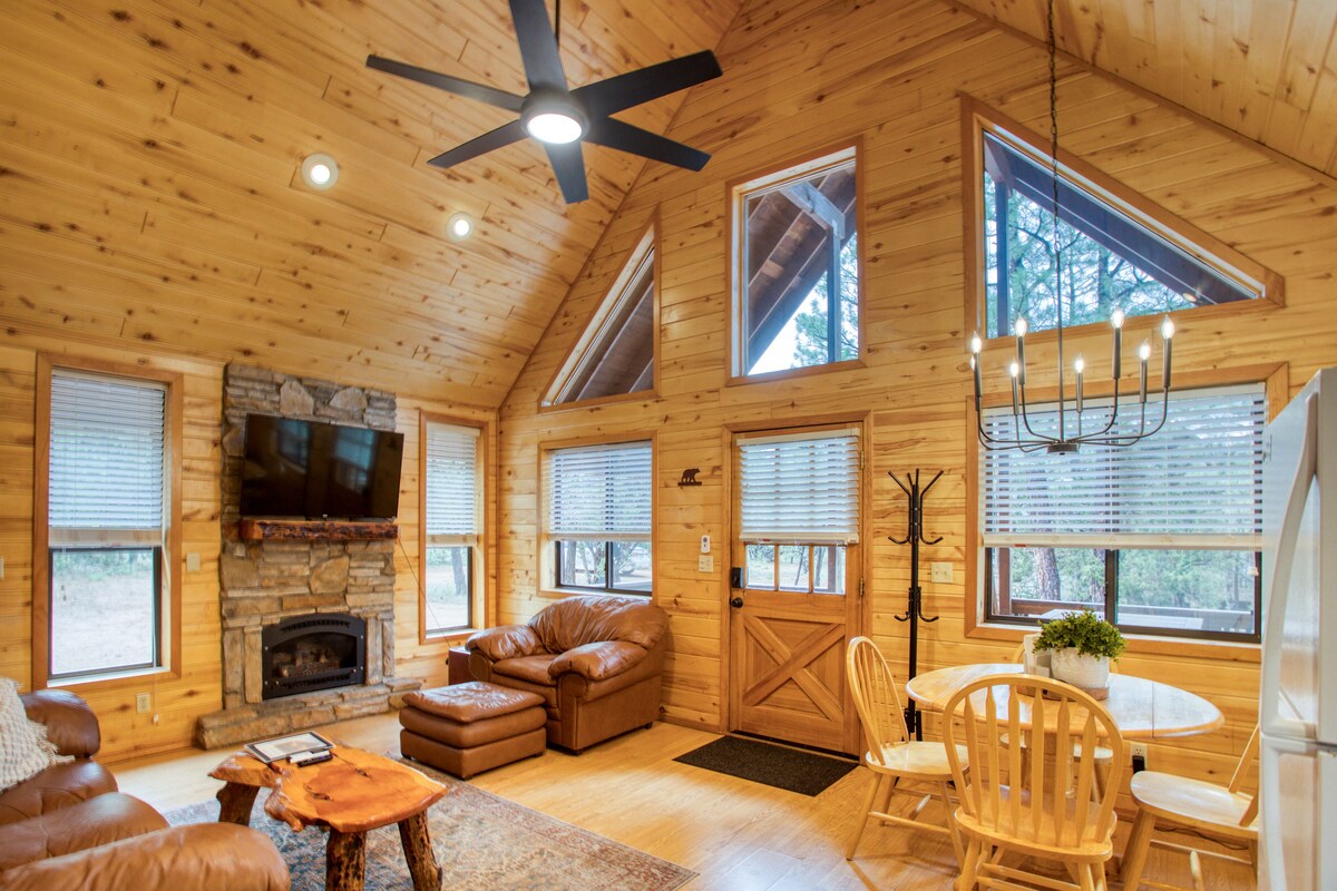 The living area features a warm wooden interior with a vaulted ceiling. A comfortable leather couch is positioned near a stone fireplace and a TV. Large windows allow natural light to fill the space, while a dining table offers seating for four.