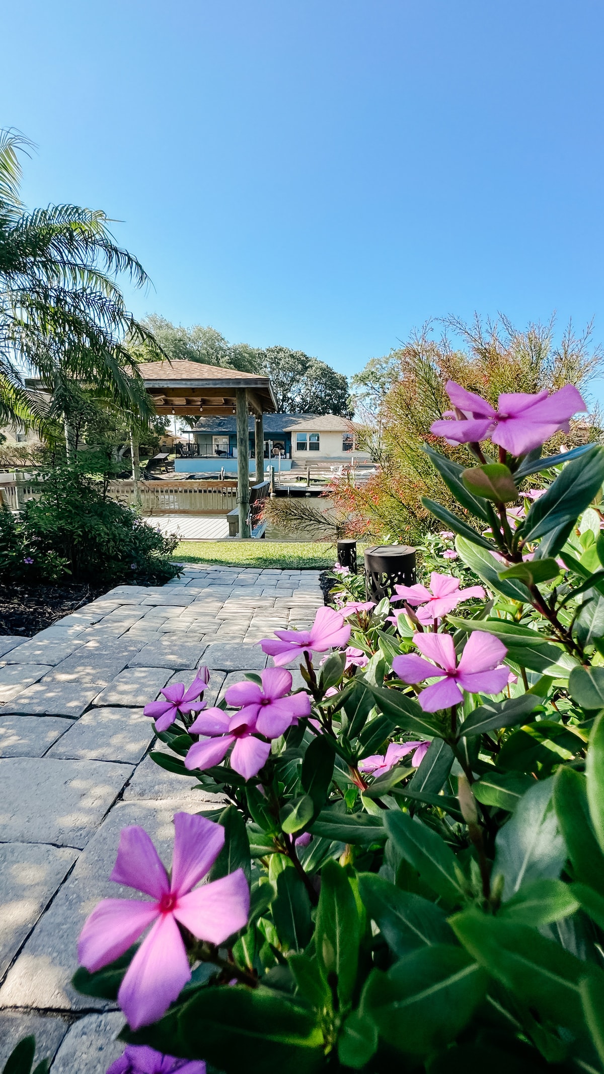 A stone pathway lined with vibrant pink flowers leads toward a shaded gazebo overlooking the tranquil canal. Lush greenery is visible along the sides, complementing the clear blue sky and creating a serene outdoor environment.