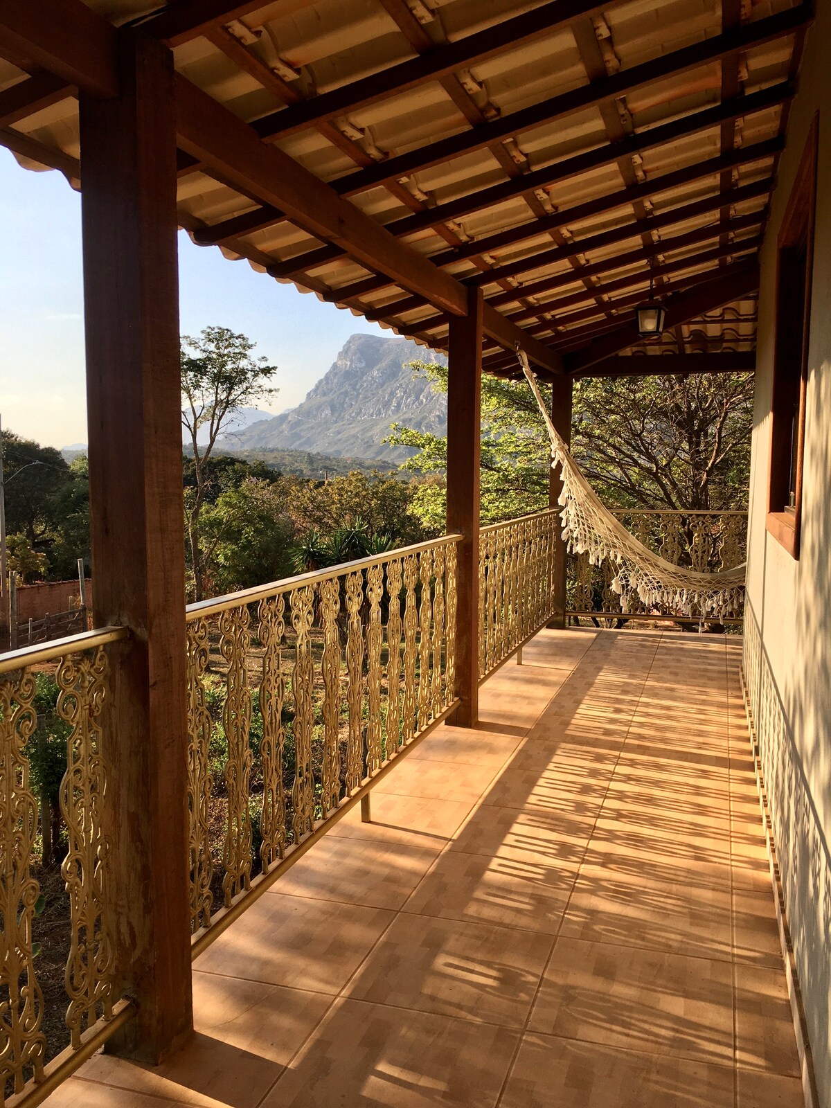 An inviting balcony is presented with a patterned railing, offering views of the surrounding greenery and distant mountains. A hammock is suspended, providing a space for relaxation. Natural light filters through, casting soft shadows on the tiled floor.