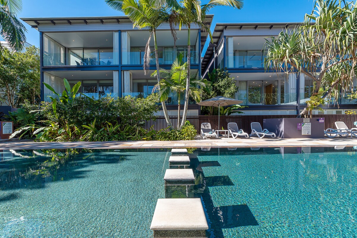 The image captures a tranquil outdoor pool area surrounded by lush tropical landscaping. Comfortable lounge chairs are positioned beside the water, with modern apartment buildings visible in the background. The calm water reflects the blue sky and greenery, creating a serene atmosphere.