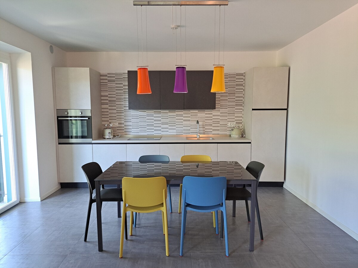 A modern kitchen area features a sleek countertop and cabinetry in light tones. A rectangular dining table is surrounded by chairs in various colors, complemented by pendant lights in vibrant hues. Two large windows provide natural light, enhancing the spacious layout.
