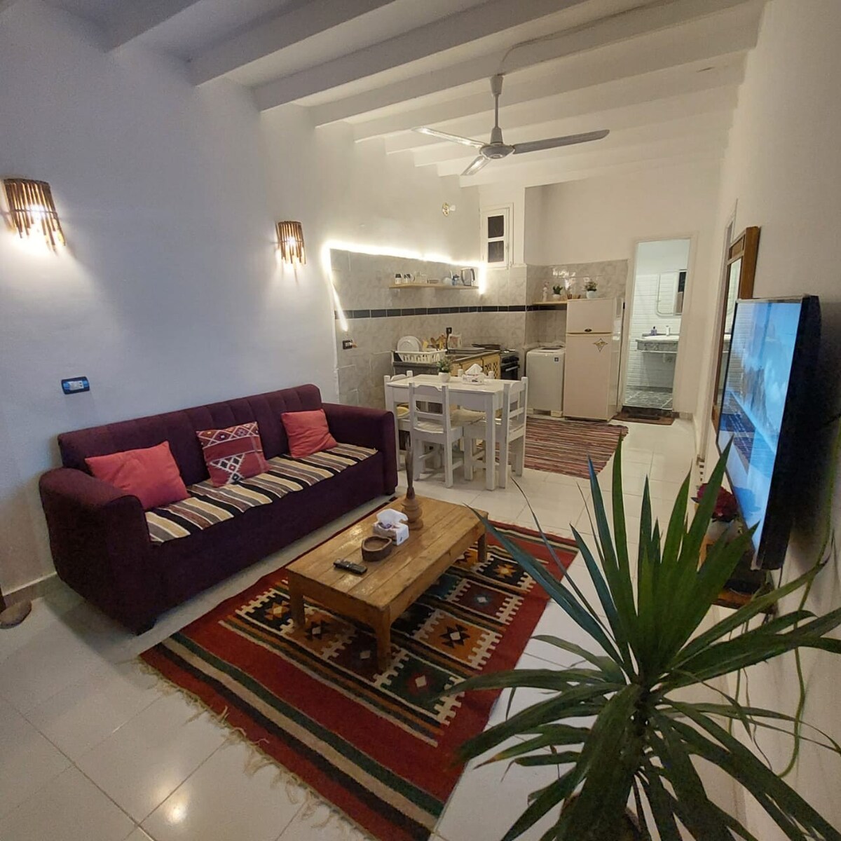 A cozy living area is presented with a dark purple sofa adorned with colorful cushions. A wooden coffee table sits center stage, surrounded by a woven rug. An open kitchen reveals modern appliances, illuminated by soft lighting, while a television is positioned on a wall opposite the seating area.