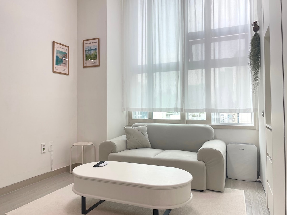 A well-lit living area features a light-colored sofa positioned beside a minimalist coffee table. Soft curtains filter natural light from the window, while framed art adds a touch of decoration to the wall. An air purifier can be seen in the corner.