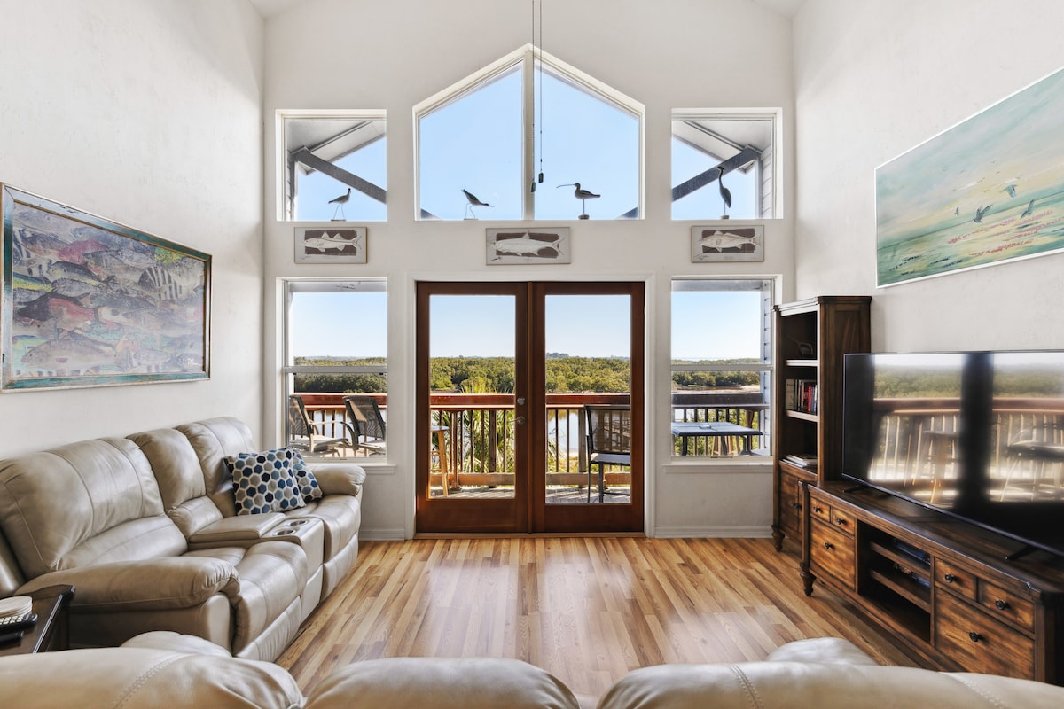 A bright living area features a large window and glass doors with stunning views of the waterfront. Two comfortable leather sofas are positioned for relaxation, complemented by wooden furniture and colorful wall art, creating a welcoming and airy atmosphere.