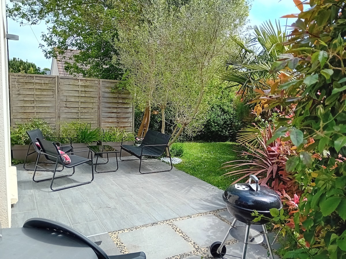 A well-maintained outdoor space features a patio with two lounge chairs and a charcoal grill. Lush greenery surrounds the area, complemented by a variety of plants and trees. The space is designed for relaxation and outdoor dining.