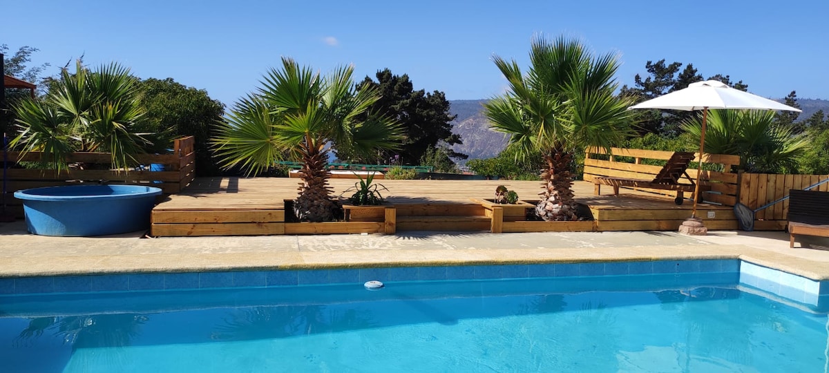 A shared swimming pool is surrounded by palm trees, wooden decking, and lounge furniture. Sun loungers are positioned under an umbrella, offering a shaded space. A decorative hot tub is also present near the pool, with a backdrop of distant mountainous scenery.
