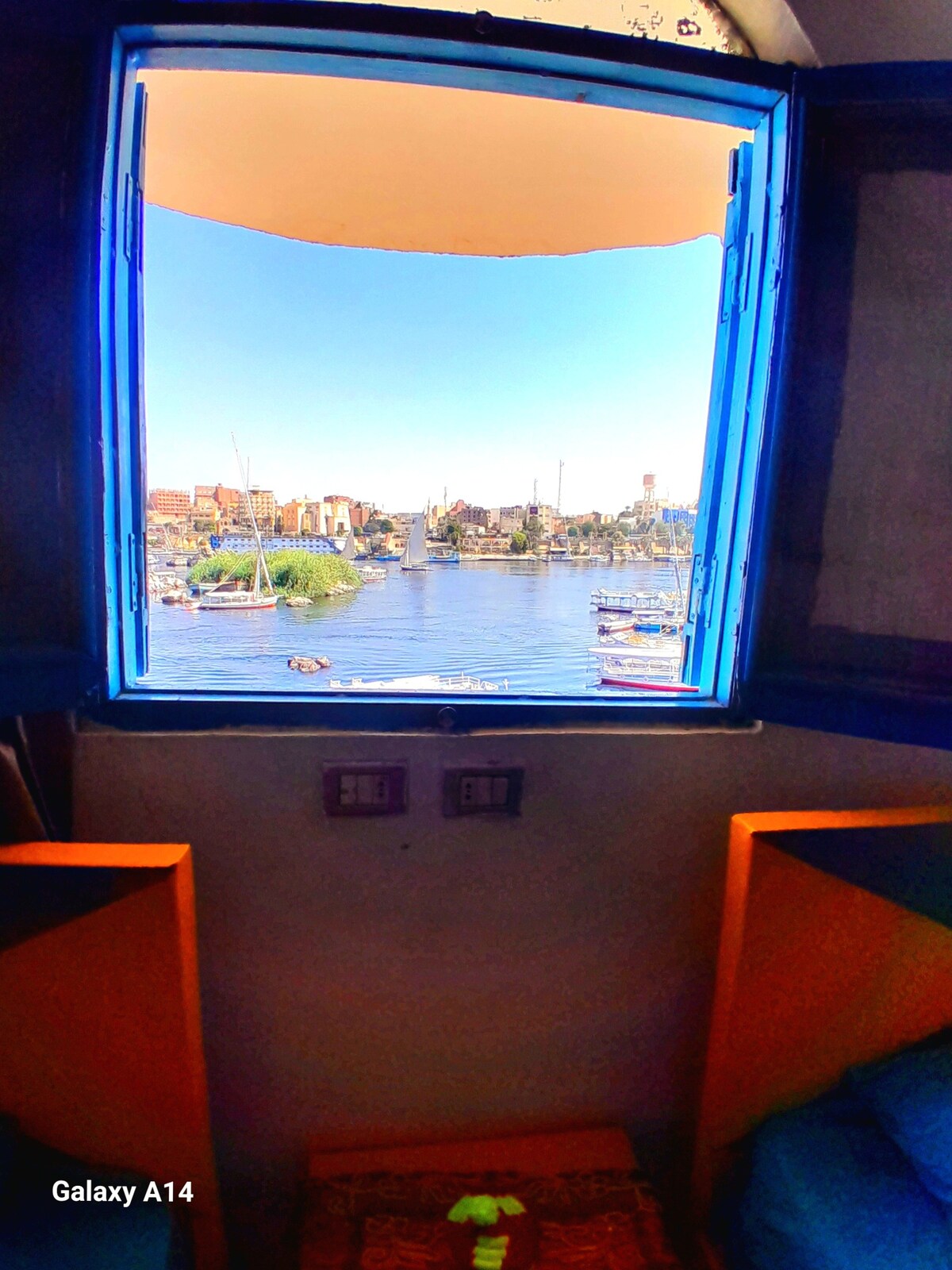 A bright window with blue frames opens to a scenic view of the river and lush greenery. The space includes two colorful seating areas placed on either side of the window, allowing for natural light to illuminate the room.