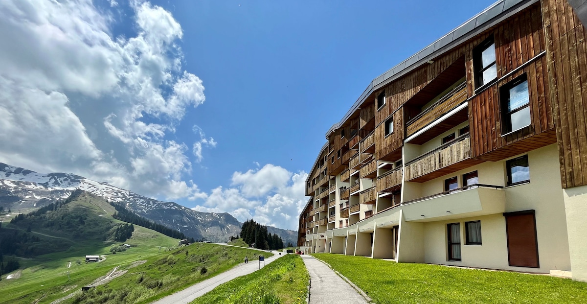 The exterior of the residence features a multi-story building designed with wooden accents and balconies. The green landscape and surrounding mountains create a scenic backdrop under a partly cloudy sky, showcasing the peaceful rural environment.
