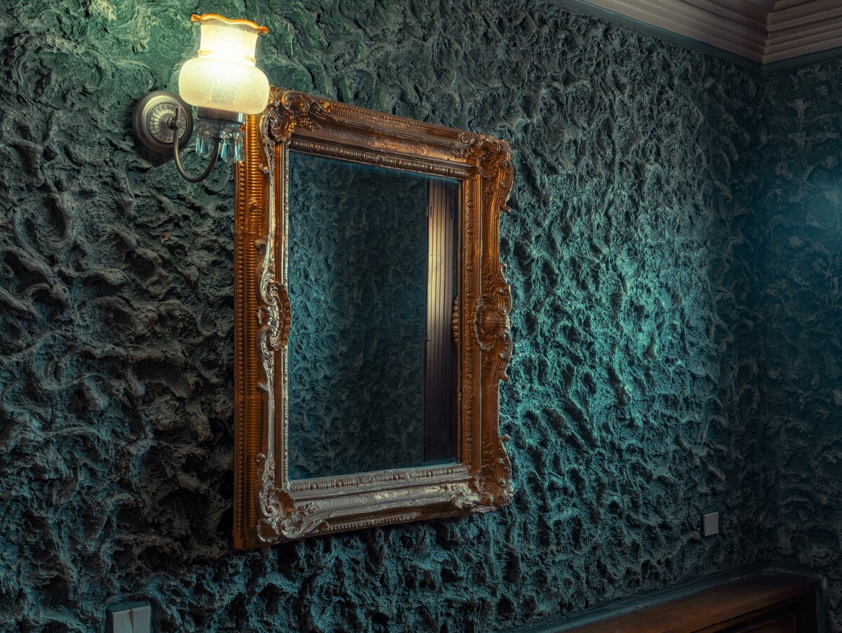 An ornate mirror with a gilded frame is mounted on a textured, dark stone wall. A vintage sconce with a frosted glass shade is positioned above the mirror, providing soft lighting that accentuates the intricate details of the frame and the unique wall texture.