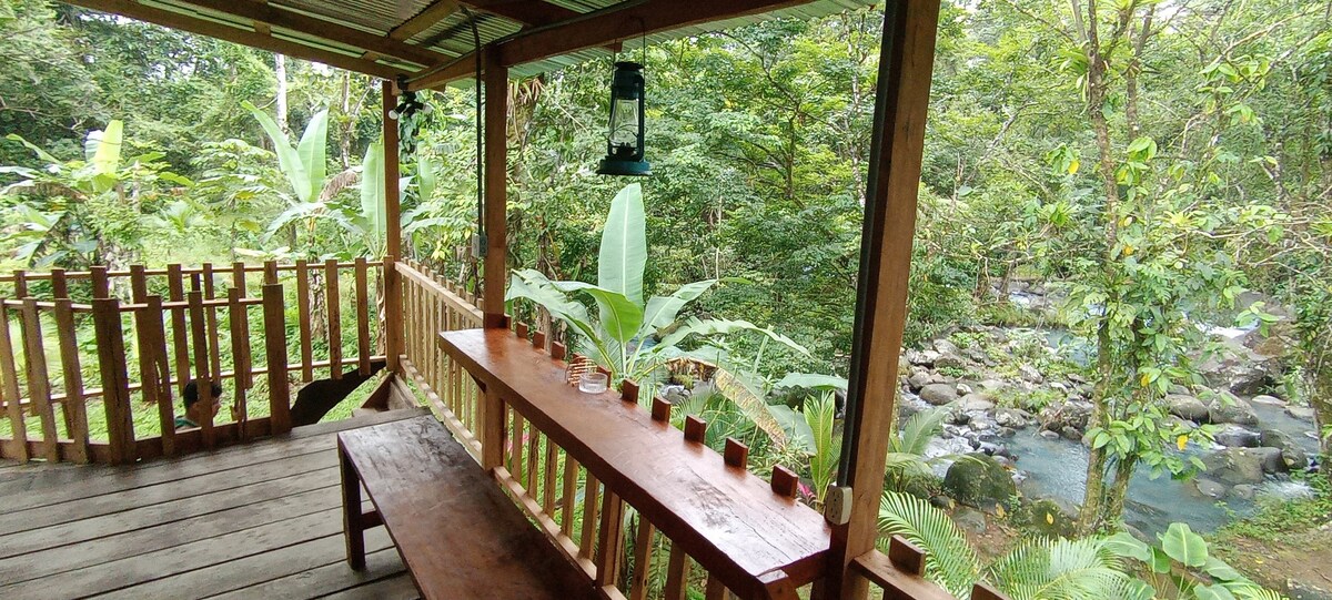 A wooden balcony features a long table and benches, surrounded by lush green foliage. A lantern hangs from the ceiling, and the serene river can be seen flowing nearby, blending with the natural landscape. The atmosphere invites relaxation and enjoyment of the scenery.