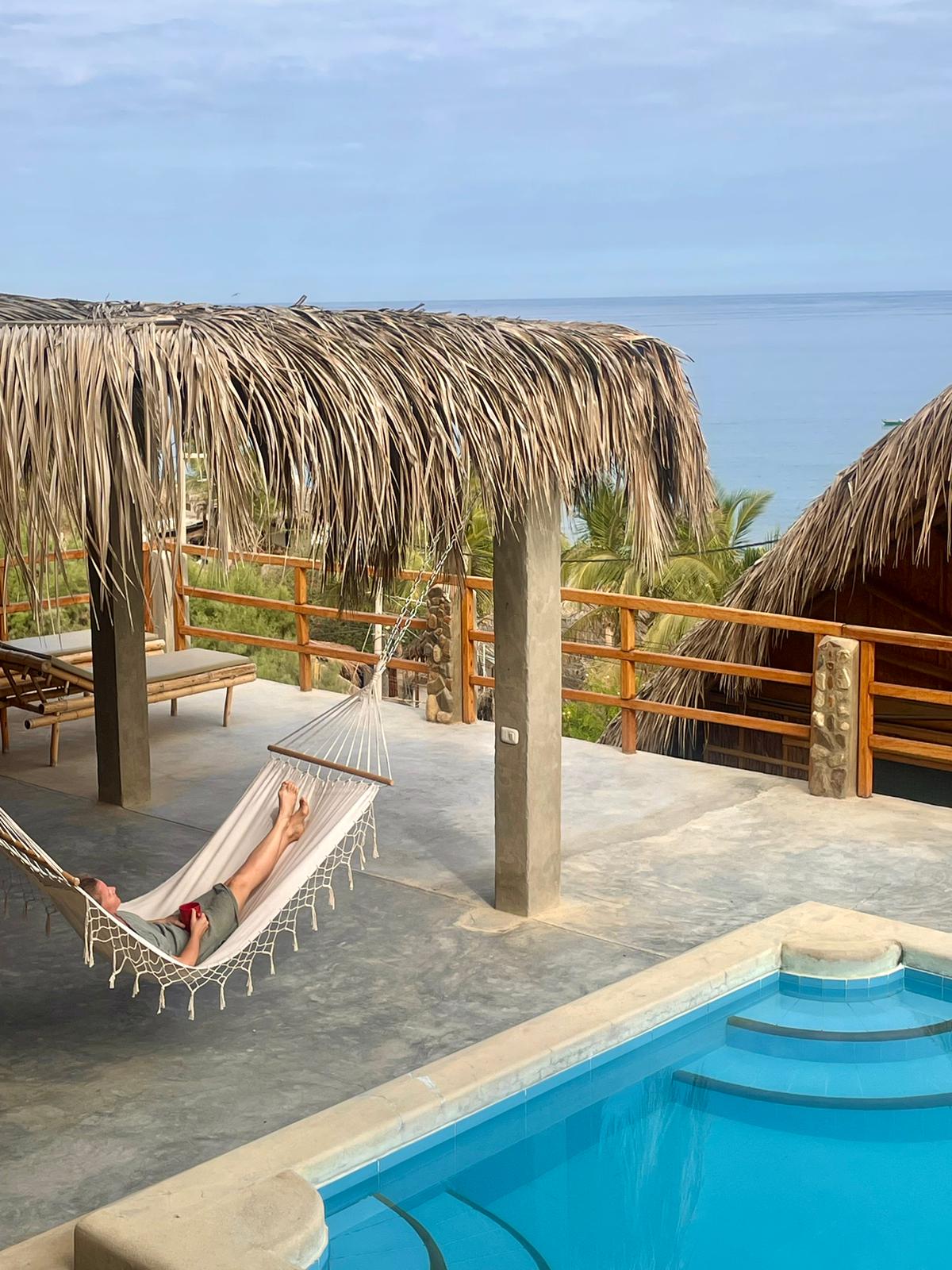 A large private terrace features a comfortable hammock suspended between two posts. A swimming pool with a gentle curve is visible in the foreground. The terrace is bordered by wooden railings, with views of the sea and surrounding palm trees in the background.