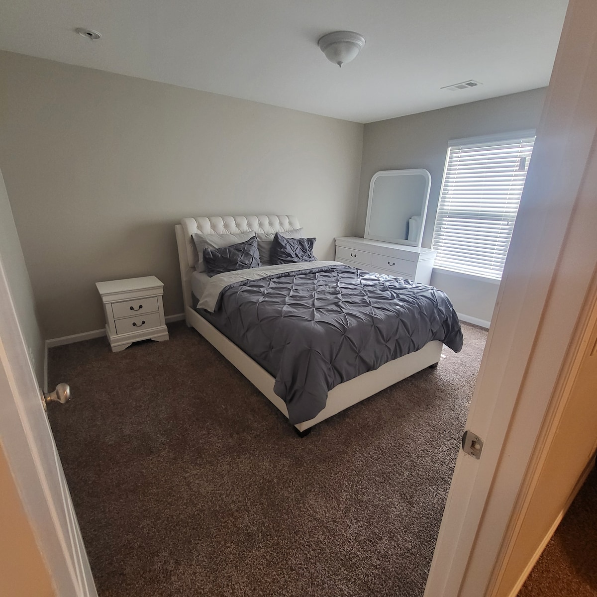 A spacious bedroom features a tufted white bed adorned with gray bedding. A matching nightstand is positioned beside the bed. Natural light enters through a window with blinds. A dresser with a mirror complements the room, accented by soft, neutral wall colors and carpeting.