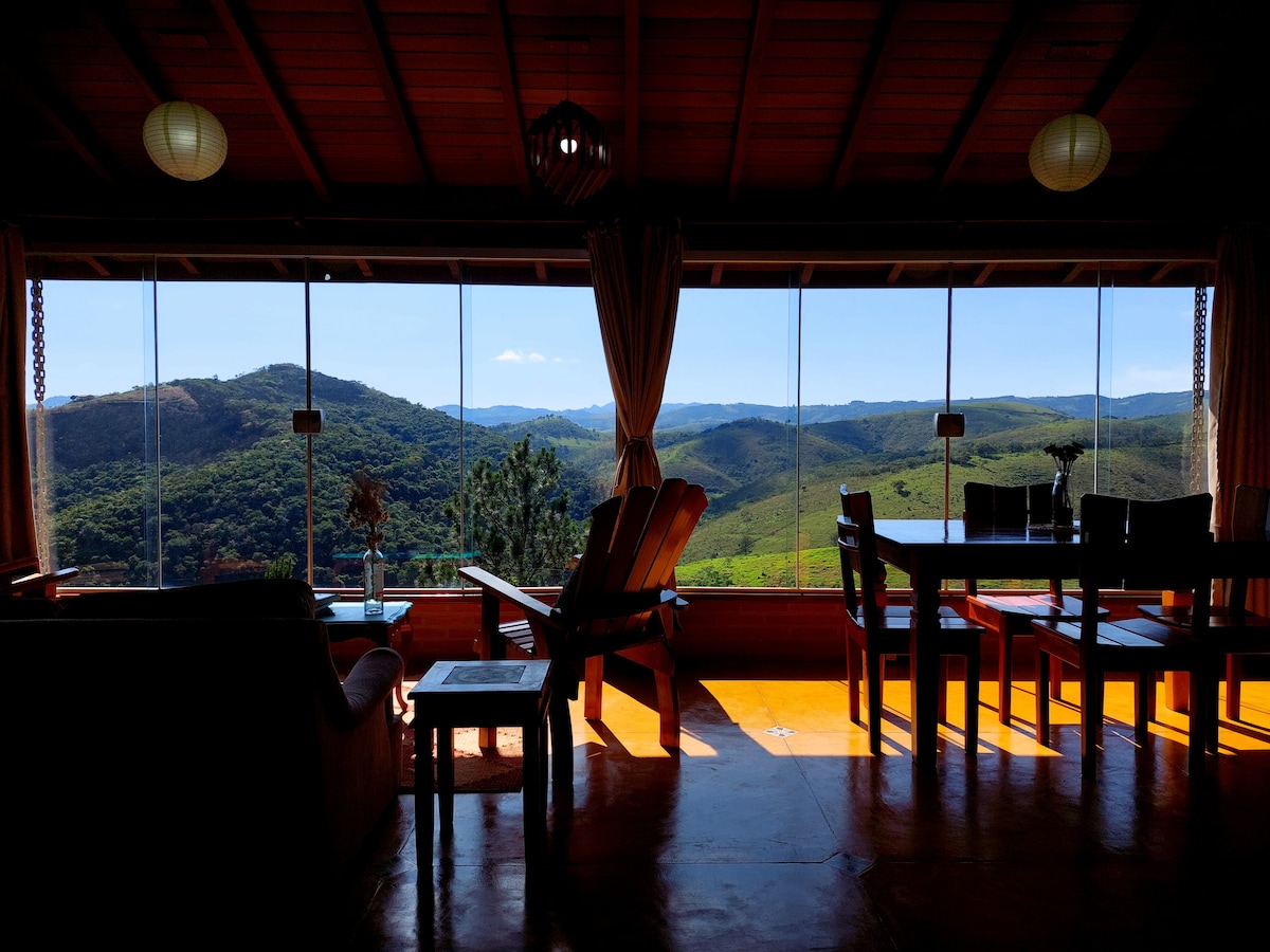 A spacious interior features large glass windows that provide breathtaking views of the green hills and mountains. Comfortable seating arrangements and a dining table are visible, illuminated by natural light. Lanterns hang from the wooden ceiling, adding an inviting touch to the space.