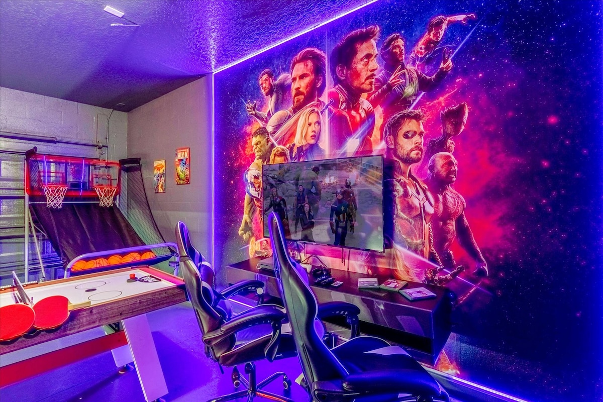 A vibrant game room features neon colored lighting and a striking mural depicting popular characters. Two ergonomic gaming chairs are positioned in front of a large screen. An air hockey table and basketball arcade game are visible, creating an inviting space for entertainment.